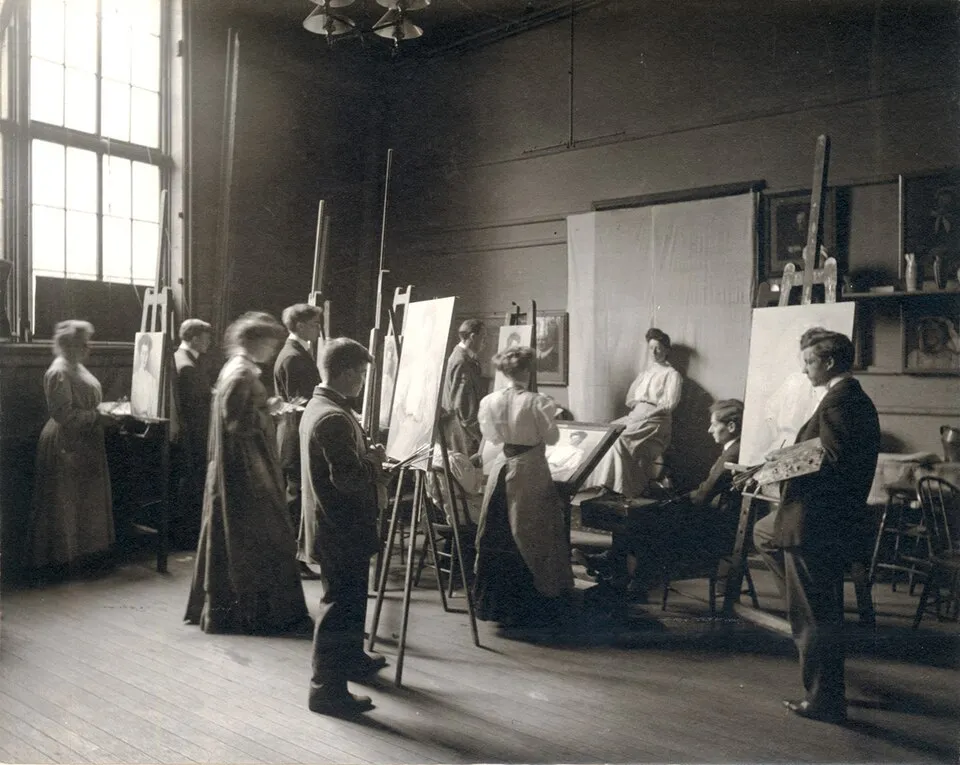 A historical photograph of a studio painting class at the Yale School of Fine Arts circa 1900-1920, showing male and female students at easels with a model, illustrating the traditional studio art class format that continues in community colleges and art schools today