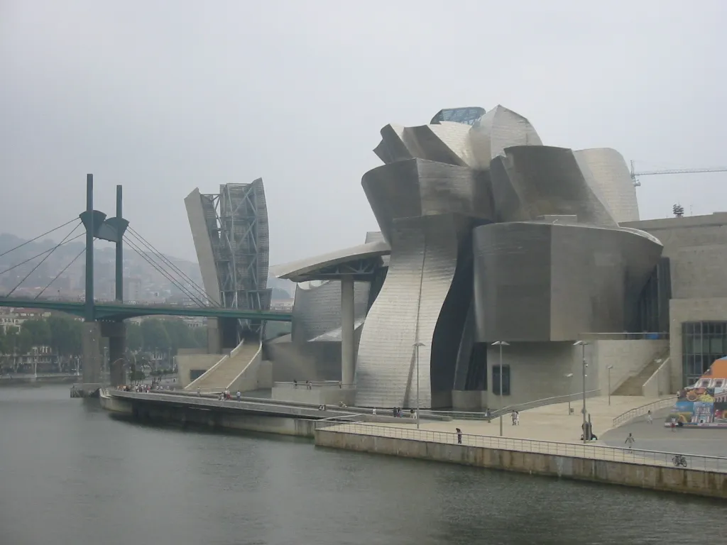 The Guggenheim Museum Bilbao (1997) by Frank Gehry, showing the titanium-clad sculptural building on the waterfront of the Nervión River in Bilbao, Spain, with its organic curving forms reflecting the sky