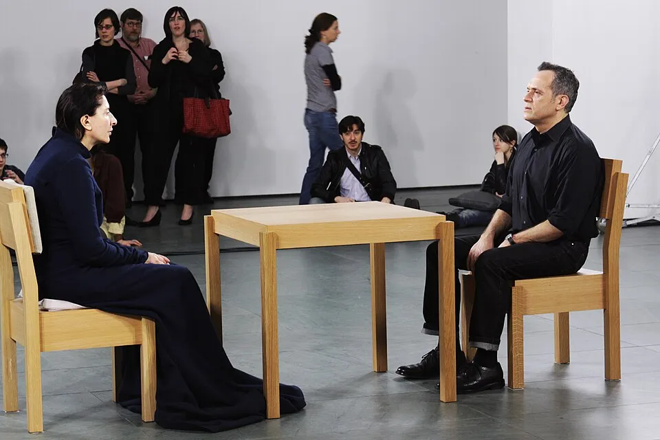 Marina Abramović sitting silently across from a museum visitor at a wooden table during 'The Artist Is Present' performance at MoMA in 2010, demonstrating the use of the body as medium in contemporary performance art