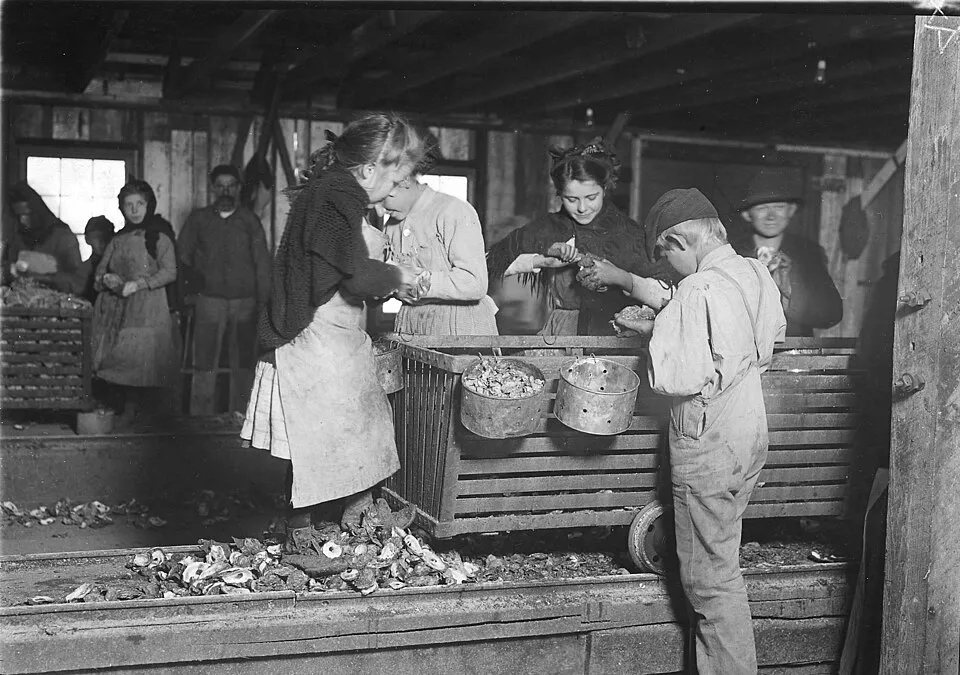 Little Lottie, a regular oyster shucker in Alabama (c. 1911) by Lewis Hine, showing a young girl standing in work clothes outside a canning facility