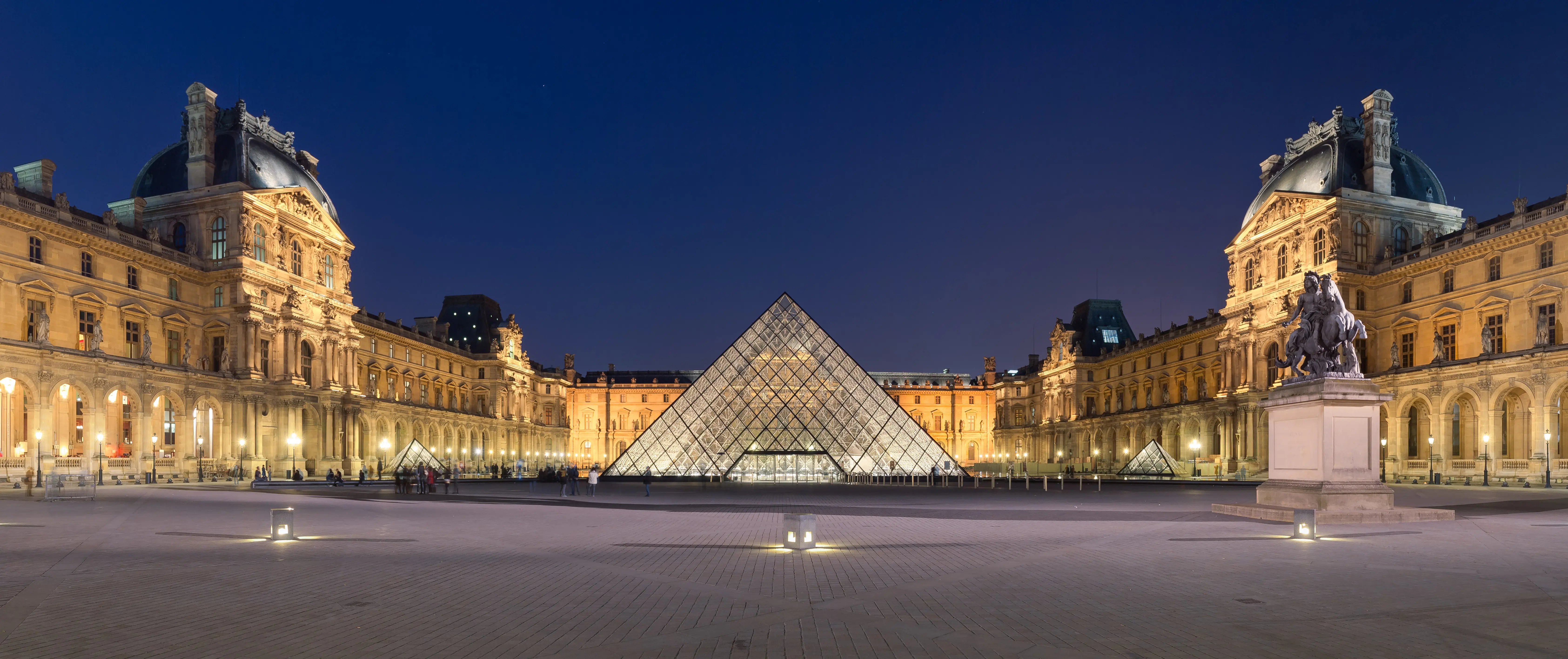 The interior of the Louvre Museum showing visitors walking through a grand gallery with paintings on the walls