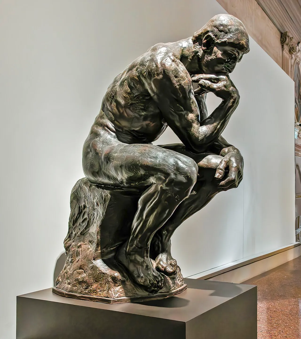 The Thinker by Auguste Rodin showing a muscular male figure seated and bent forward in deep contemplation, with chin resting on hand, cast in dark bronze against a clear sky