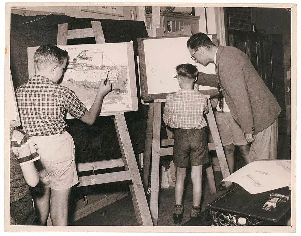 A group of children in an art class, painting at tables with brushes and colorful paints, demonstrating the hands-on, process-oriented approach to early art education that research shows develops creativity, confidence, and cognitive skills